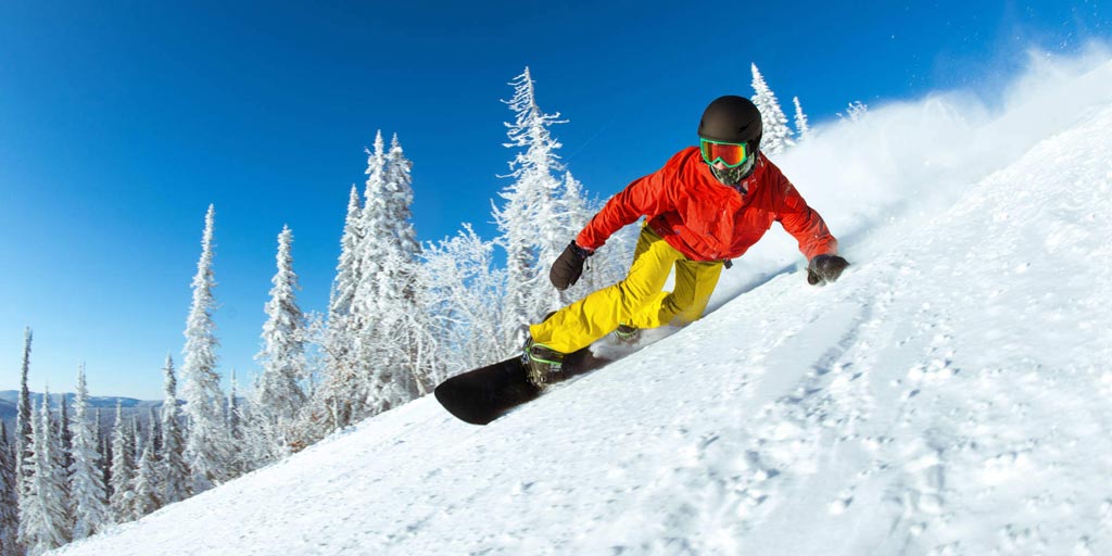 A snowboarder wearing bright colours turning on a snowy mountainside against a blue sky and snowy trees background